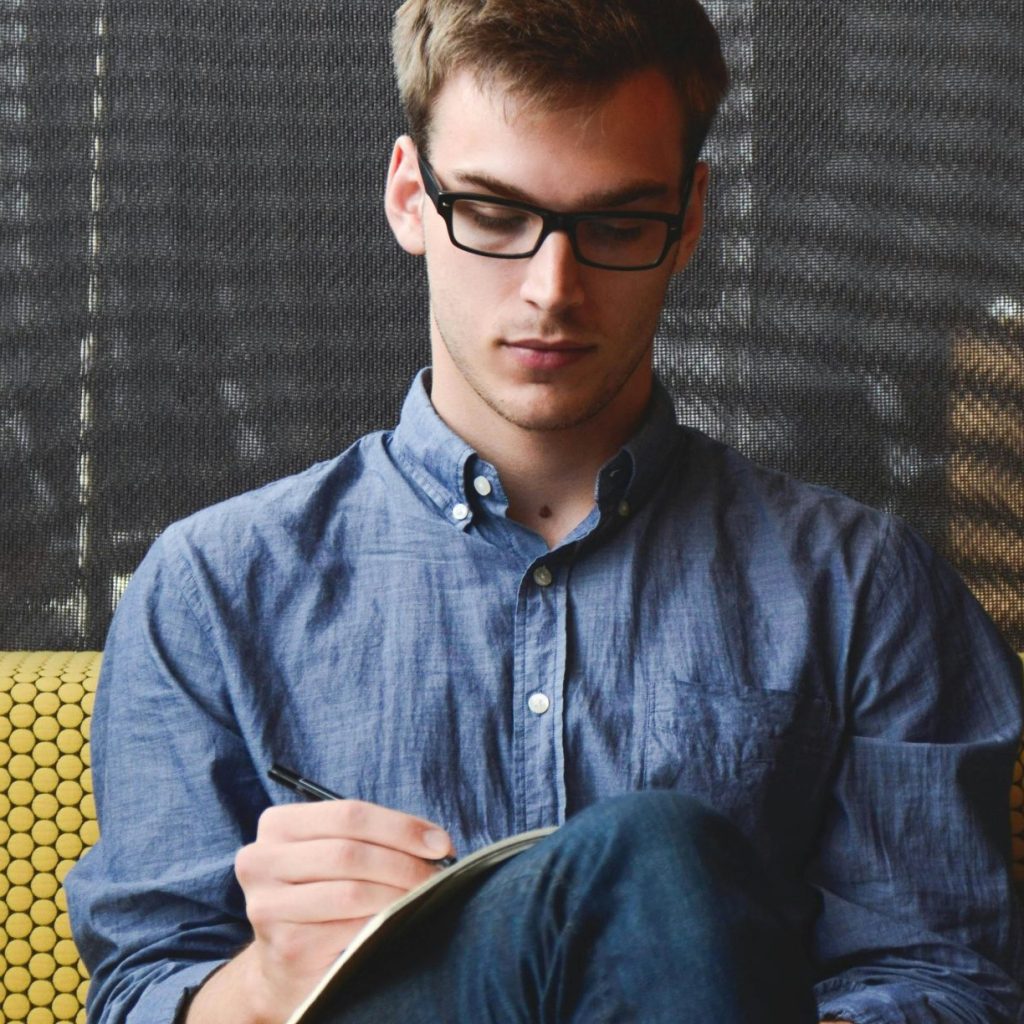 A young man in glasses writes in a notebook while sitting on a stylish couch indoors.